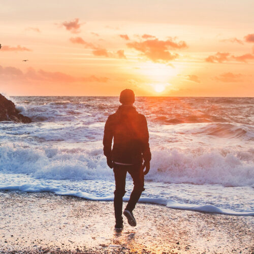 Mann geht am Strand Richtung Sonnenuntergang